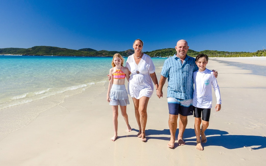 Family walking on Whitehaven Beach, Airlie Beach, with clear blue water and green hills in the background.