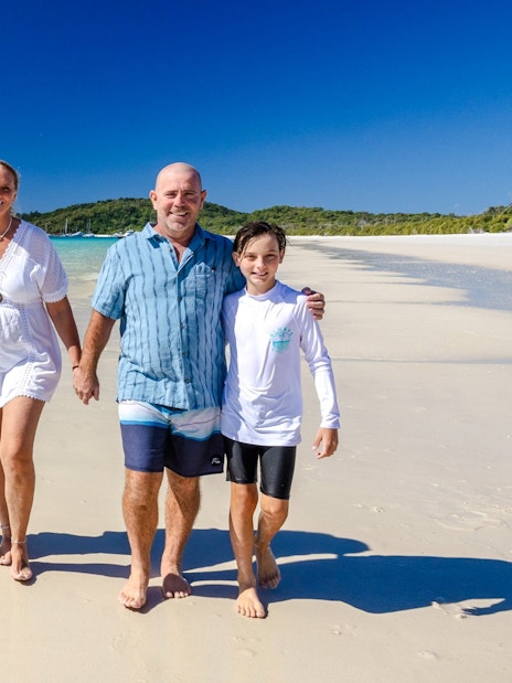 Family walking on Whitehaven Beach, Airlie Beach, with clear blue water and green hills in the background.