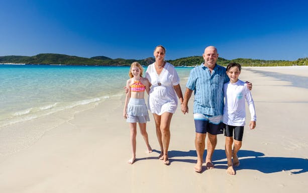 Family walking on Whitehaven Beach, Airlie Beach, with clear blue water and green hills in the background.