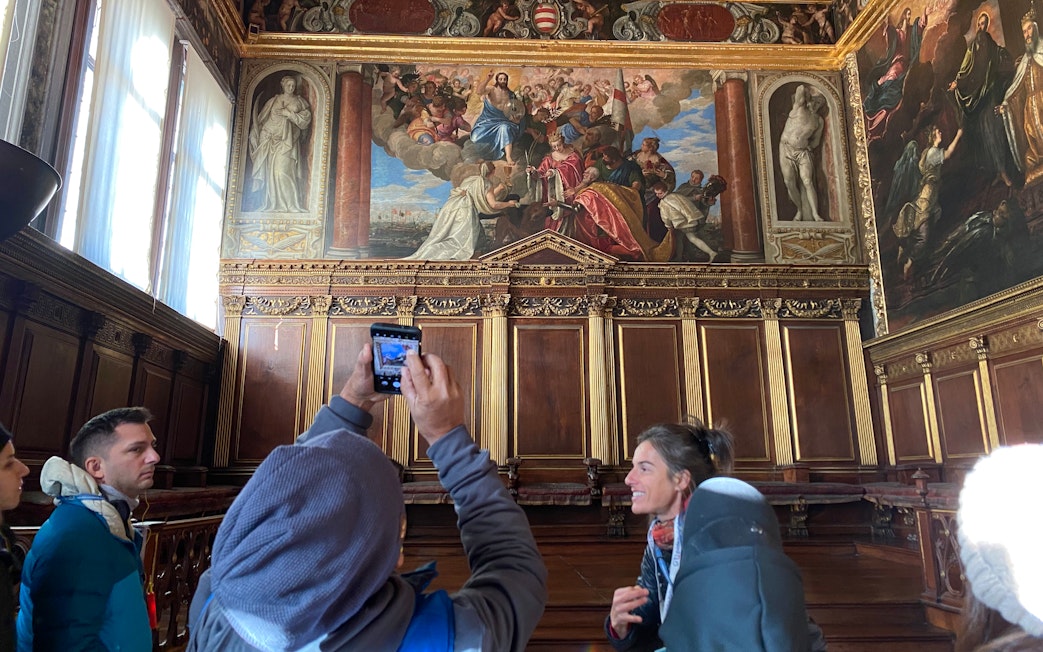 Tour group inside Doge's Palace viewing ornate frescoes and wood paneling.