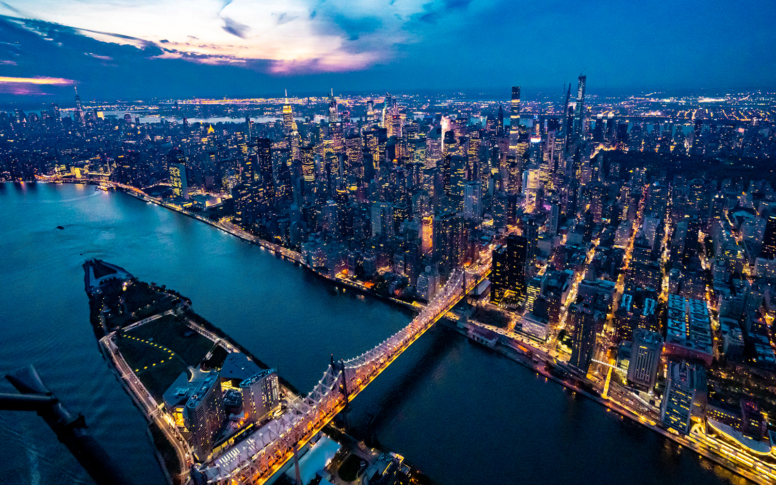 Aerial view of New York City skyline at dusk, featuring illuminated skyscrapers and East River.