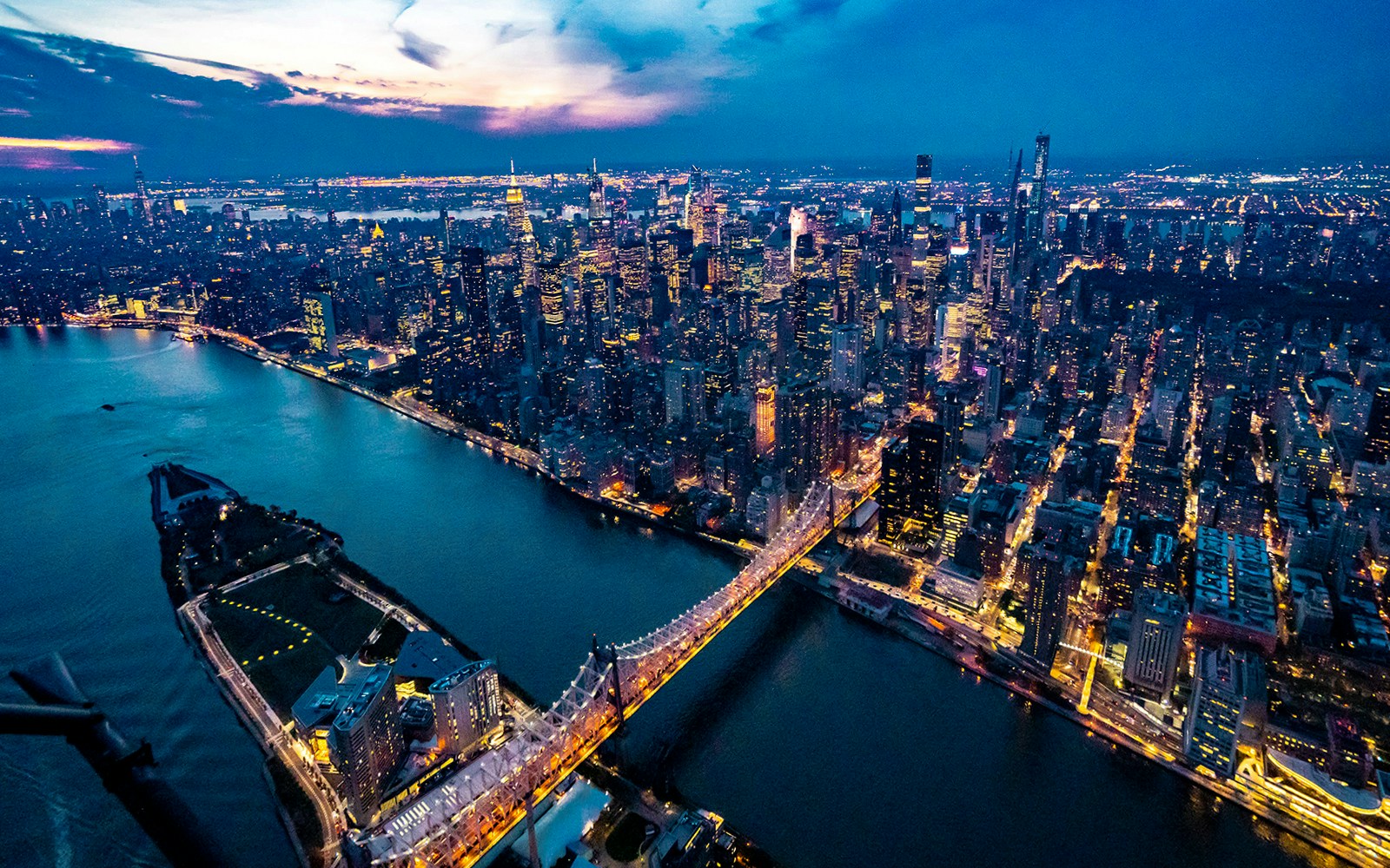 Aerial view of New York City skyline at dusk, featuring illuminated skyscrapers and East River.