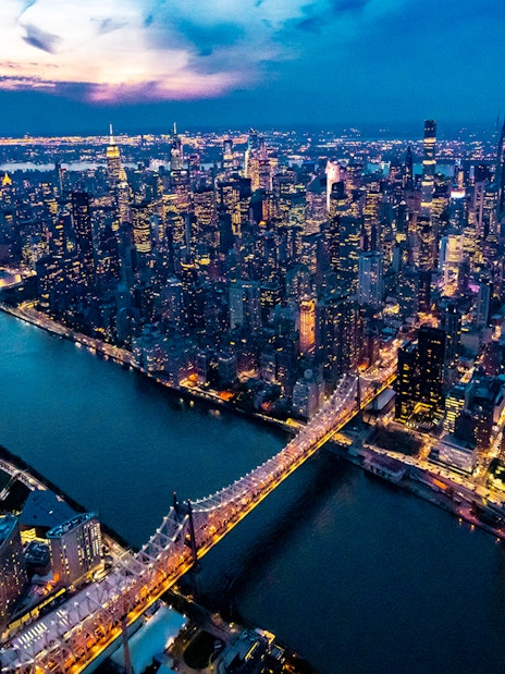 Aerial view of New York City skyline at dusk, featuring illuminated skyscrapers and East River.