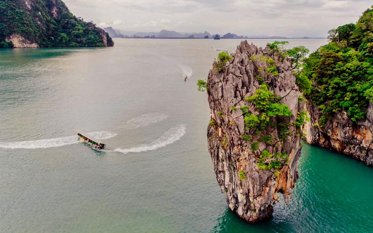 Towering limestone cliff with lush greenery in emerald waters, boat passing by.
