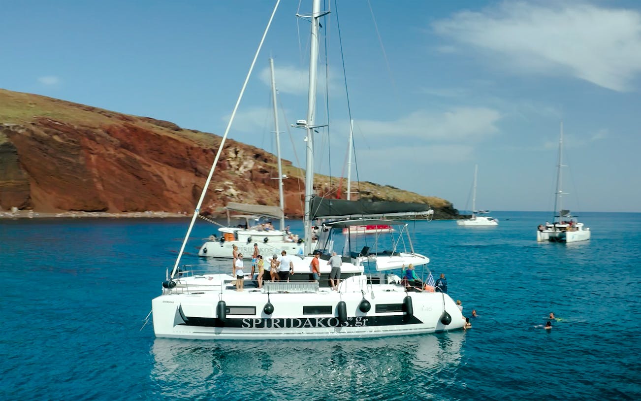 Catamaran cruise near Santorini's red cliffs with people swimming nearby.