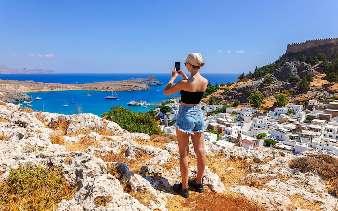 Tourist photographing the Acropolis of Lindos with a view of the Aegean Sea and Lindos village.