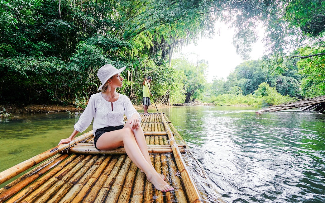 Woman on bamboo raft in lush tropical river setting.