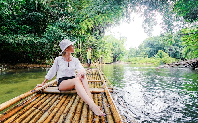 Woman on bamboo raft in lush tropical river setting.
