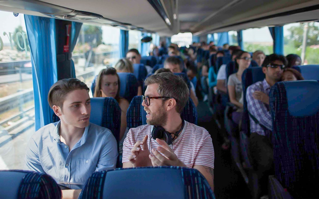 Passengers conversing on a tour bus with blue seats and curtains.