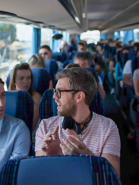 Passengers conversing on a tour bus with blue seats and curtains.