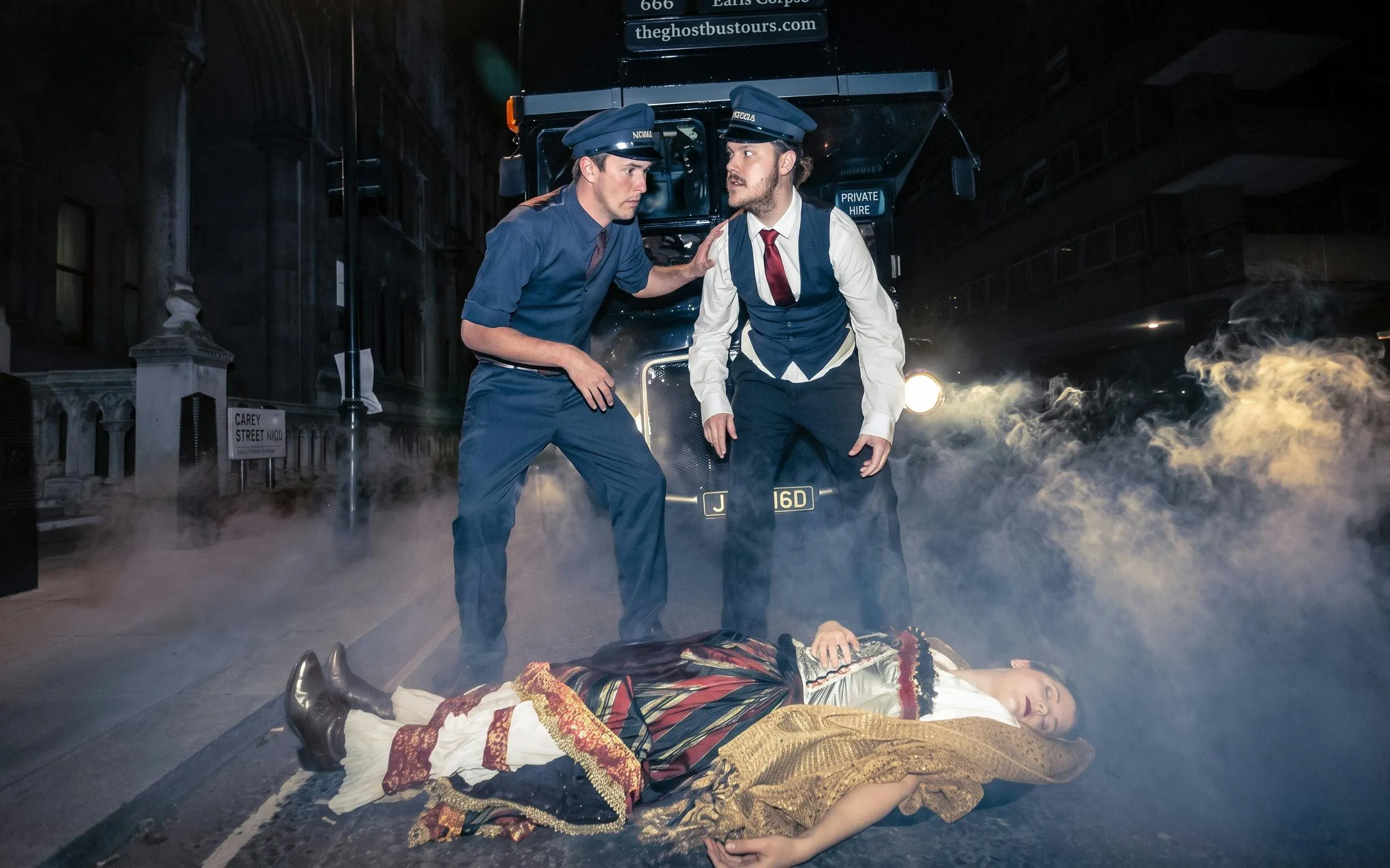 Two actors in vintage uniforms beside a bus with a costumed figure on the ground, London ghost bus tour.