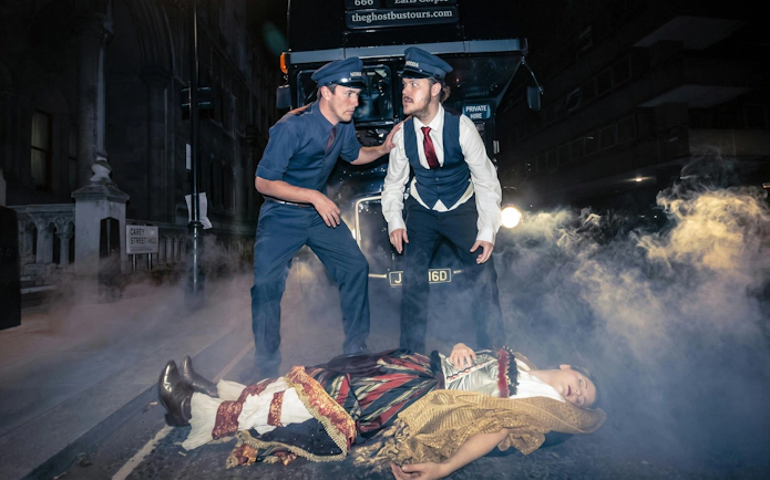 Two actors in vintage uniforms beside a bus with a costumed figure on the ground, London ghost bus tour.