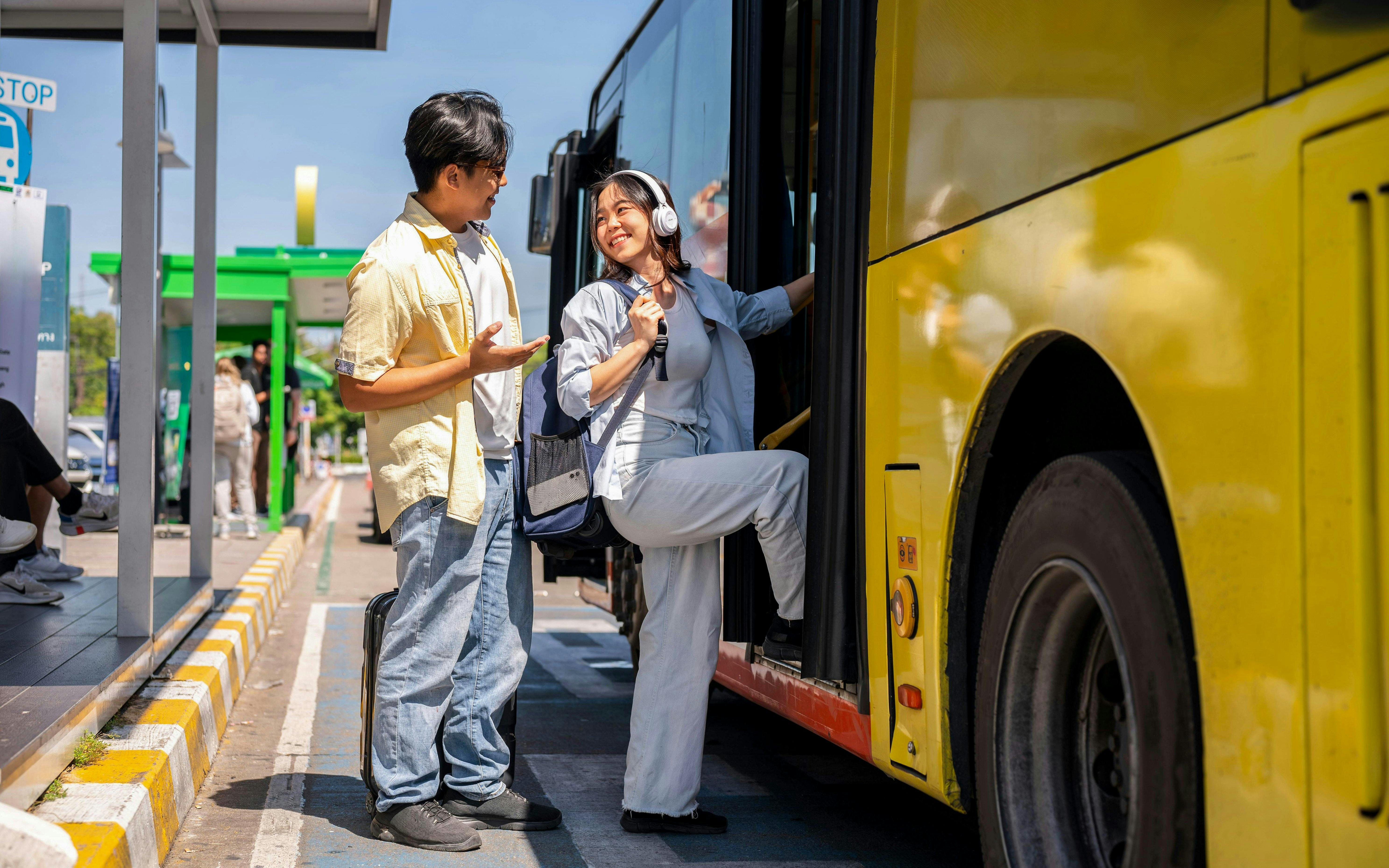 Asian tourist couple conversing at shuttle bus stop, woman boarding yellow bus.