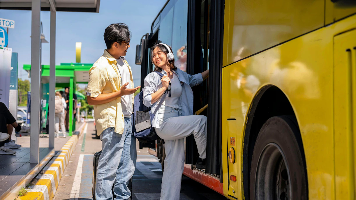 Asian tourist couple conversing at shuttle bus stop, woman boarding yellow bus.