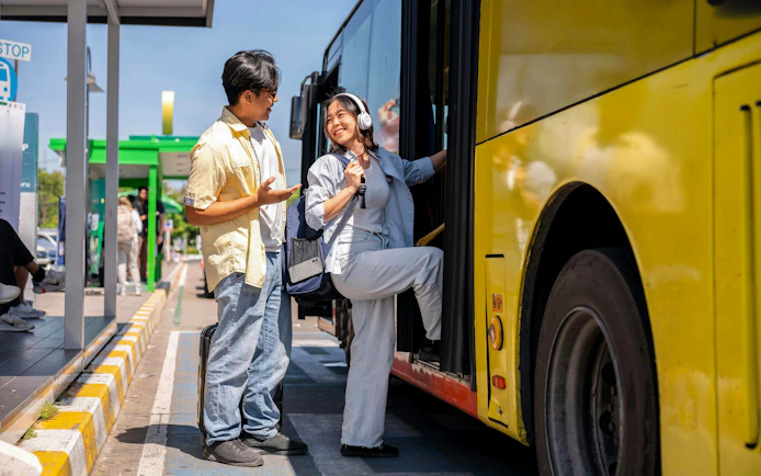 Asian tourist couple conversing at shuttle bus stop, woman boarding yellow bus.
