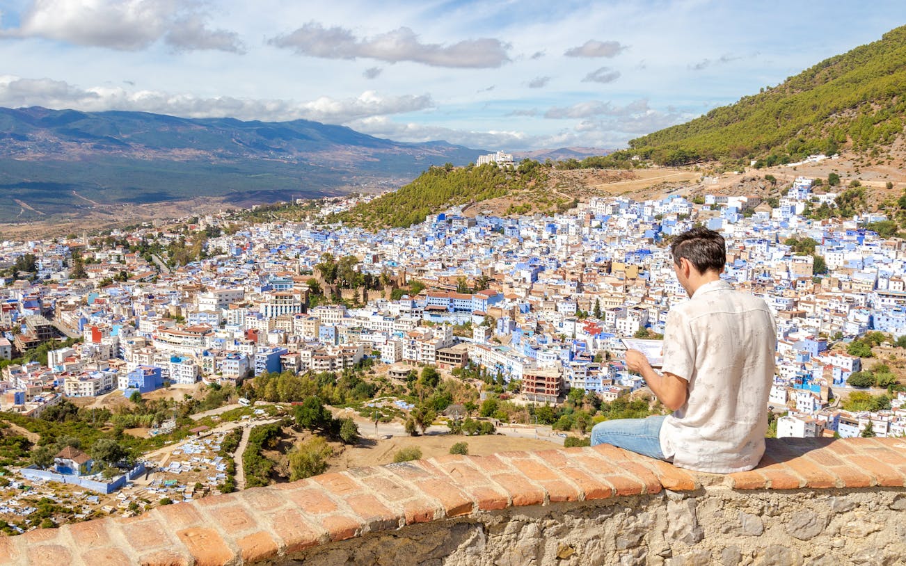 Man sitting on a wall overlooking the blue buildings of Chefchaouen, Morocco.