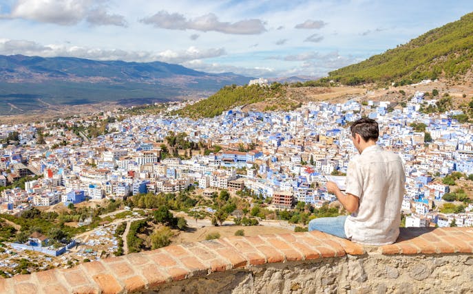 Man sitting on a wall overlooking the blue buildings of Chefchaouen, Morocco.