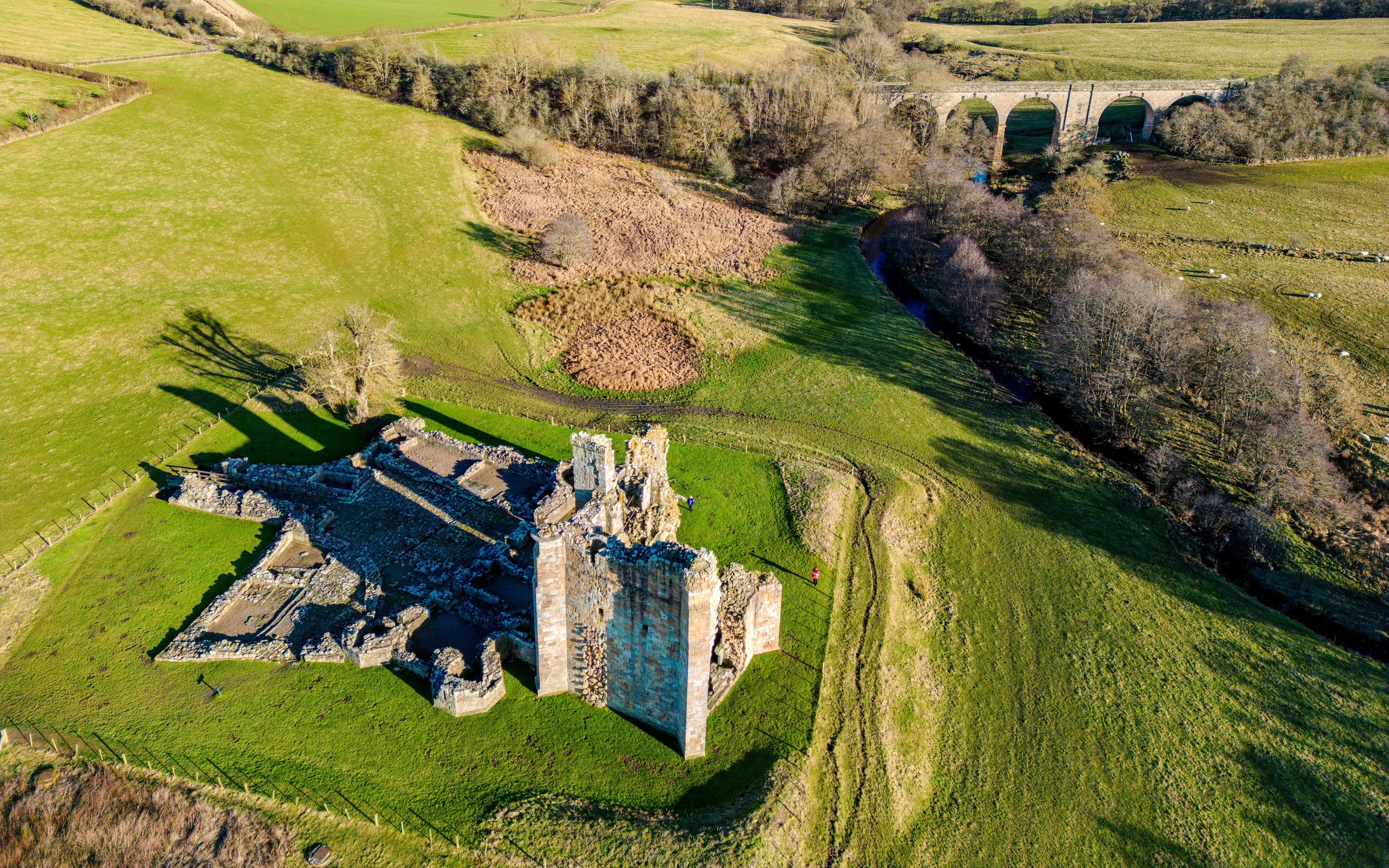 Ruins of Alnwick Castle surrounded by green fields and a stone viaduct in the distance.