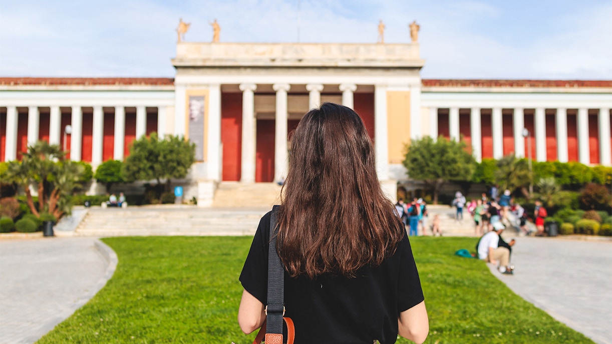 Woman staning in front of National Archaeological Museum entrance facade, Athens, Greece, showcasing neoclassical architecture.