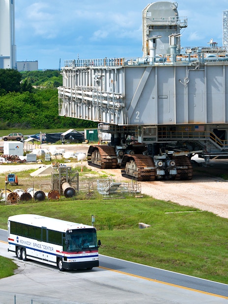 Kennedy Space Center bus near a large crawler-transporter with a launch pad in the background.