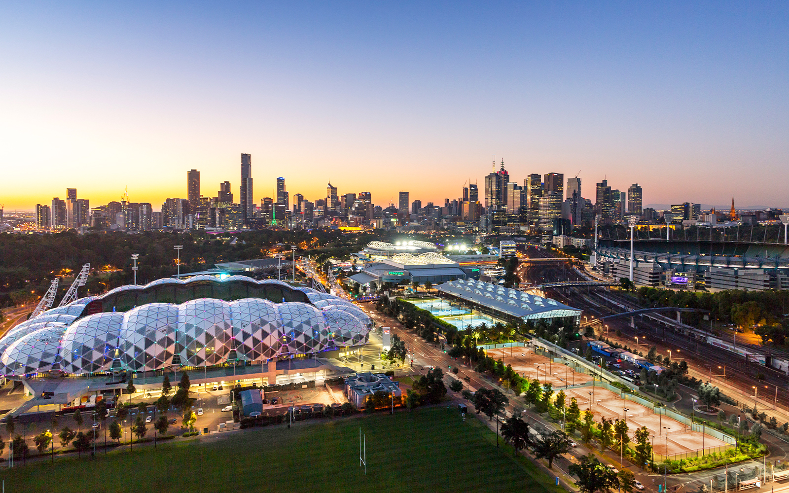 Melbourne skyline with sports venues at sunset, featuring AAMI Park and Melbourne Cricket Ground.