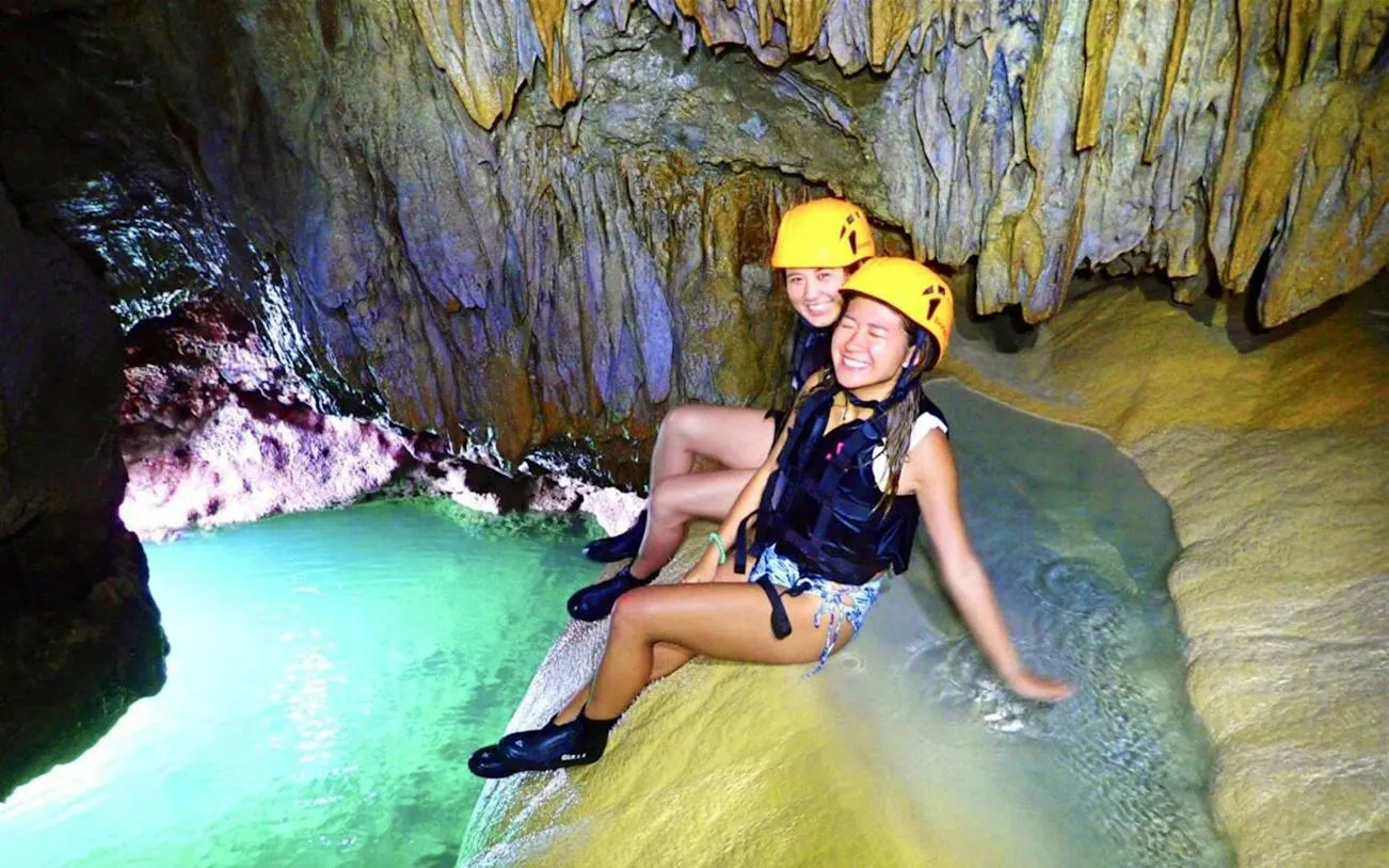 Visitors exploring Pumpkin Cave in Miyako, Okinawa, wearing helmets and life vests near a turquoise pool.