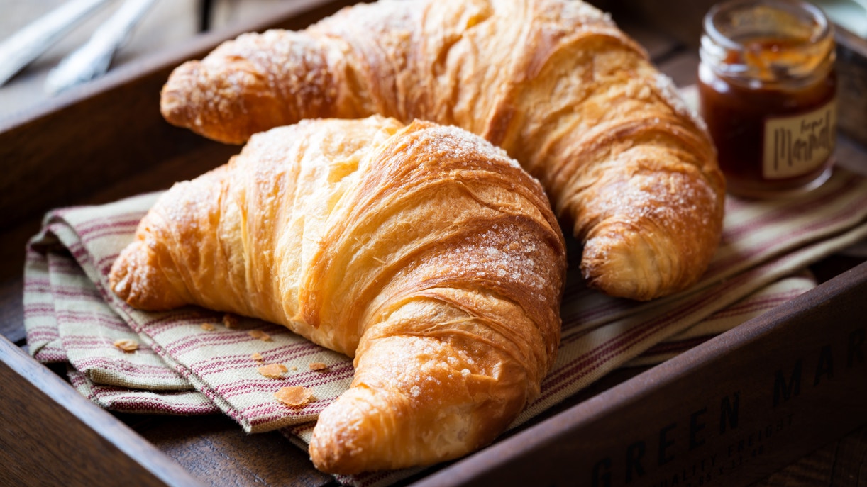 Two croissants on a striped cloth in a wooden tray with a jar of jam nearby.