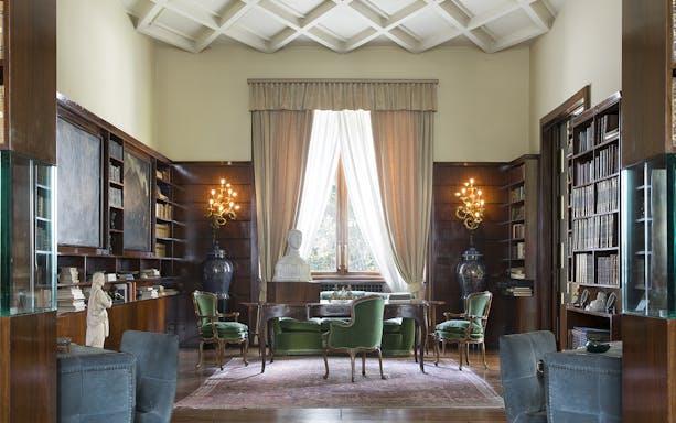 Elegant study room in a Milan house museum with bookshelves, green chairs, and a bust on a desk.