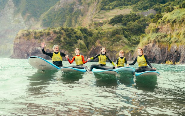 Family holding hands on surfboards during a surf lesson near rocky cliffs.