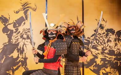 Tourists in traditional Samurai armor holding swords in front of a Japanese mural.