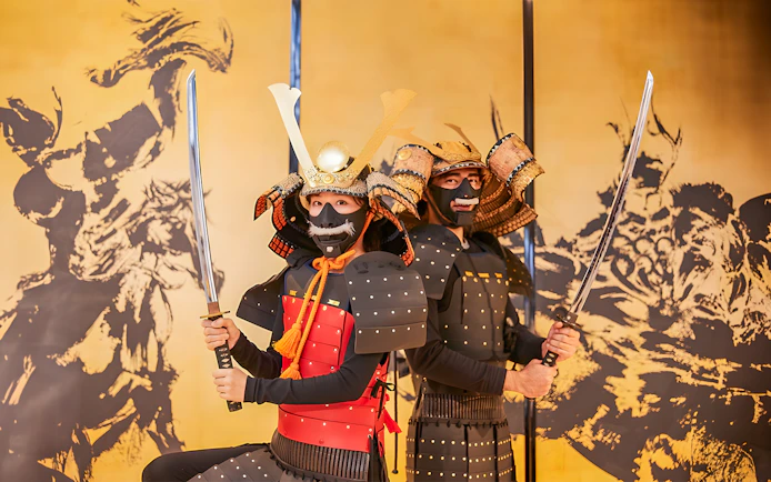 Tourists in traditional Samurai armor holding swords in front of a Japanese mural.