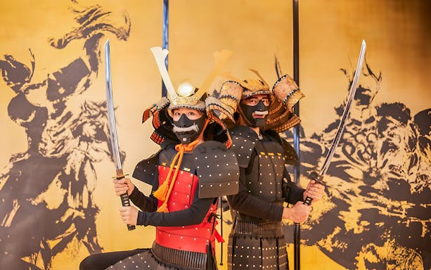Tourists in traditional Samurai armor holding swords in front of a Japanese mural.