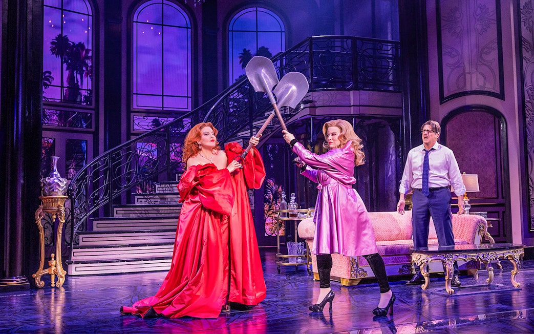 Two women in elegant dresses duel with shovels on a grand staircase set, "Death Becomes Her" theme.