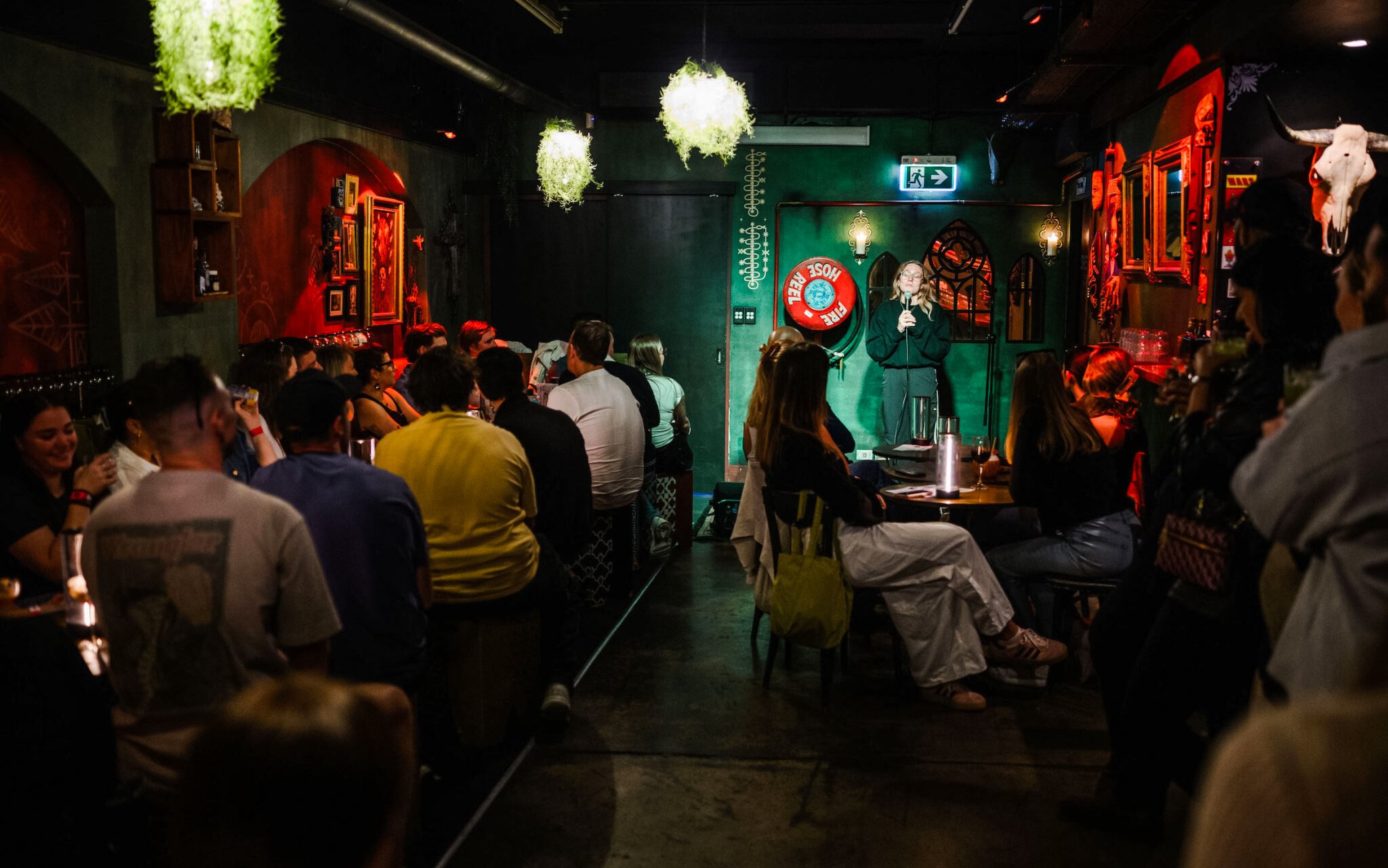Comedian performing at Sydney Comedy Festival in a dimly lit venue with audience seated.