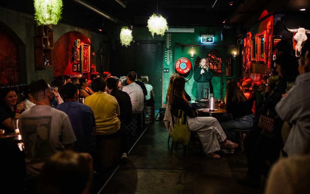 Comedian performing at Sydney Comedy Festival in a dimly lit venue with audience seated.