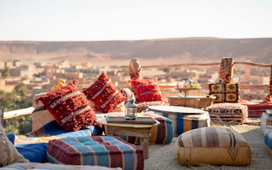 Old lamp on table with colorful cushions at rooftop restaurant overlooking desert town.