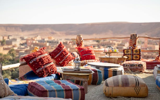 Old lamp on table with colorful cushions at rooftop restaurant overlooking desert town.