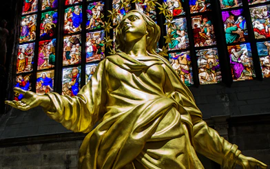 Golden statue inside Milan Cathedral with colorful stained glass windows in the background.