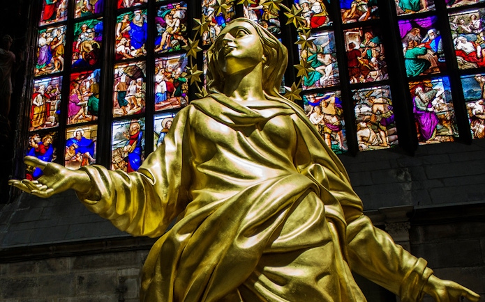 Golden statue inside Milan Cathedral with colorful stained glass windows in the background.
