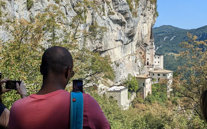 Visitors photographing Madonna della Corona Sanctuary nestled in cliffs, Italy.