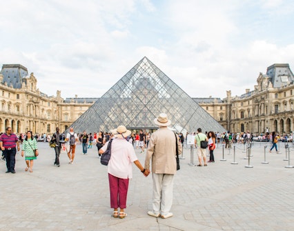 Old couple holding hands in front of the Louvre Pyramid, Paris.