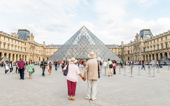 Old couple holding hands in front of the Louvre Pyramid, Paris.