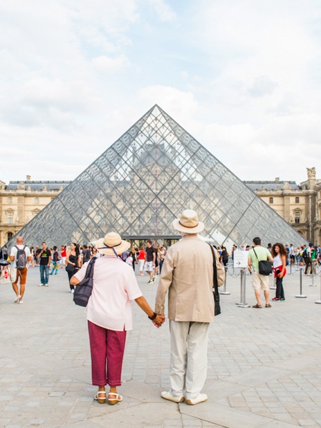 Old couple holding hands in front of the Louvre Pyramid, Paris.