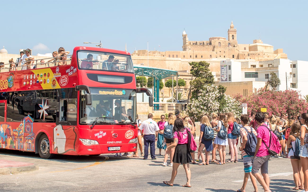 Open-top tour bus in Gozo with tourists boarding near historic buildings.