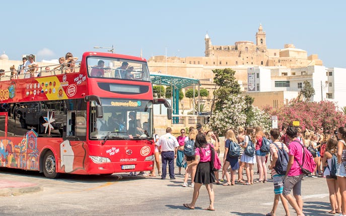 Open-top tour bus in Gozo with tourists boarding near historic buildings.