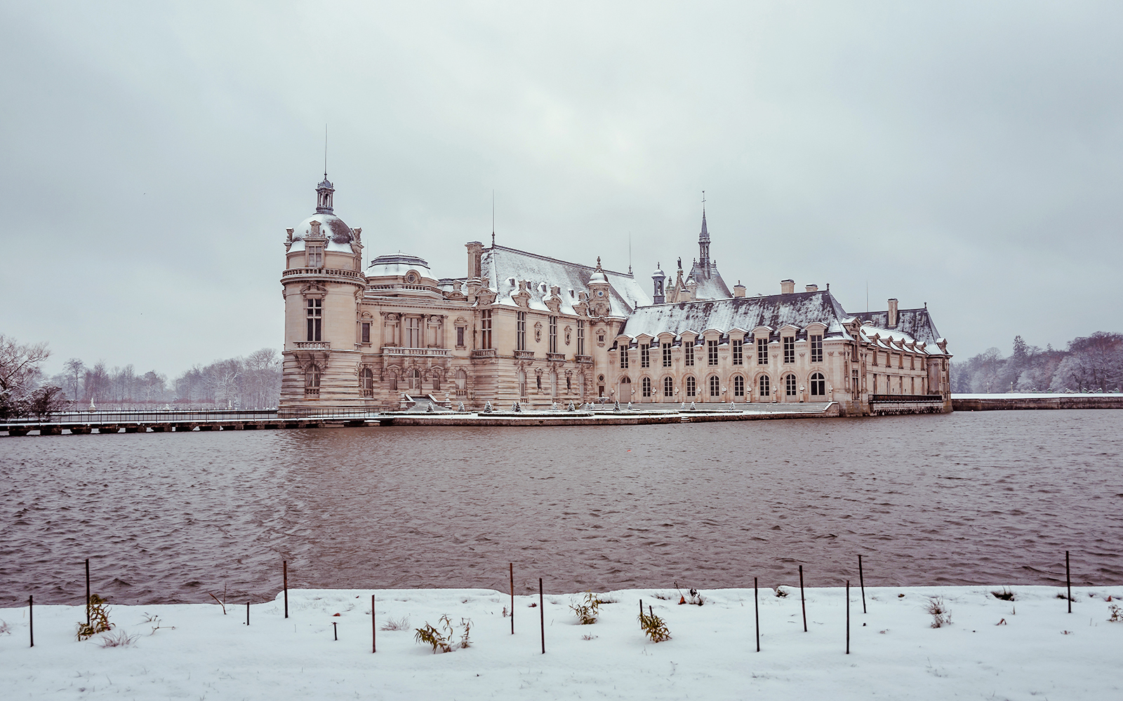 Chateau of Chantilly in winter with snow-covered grounds and surrounding water.