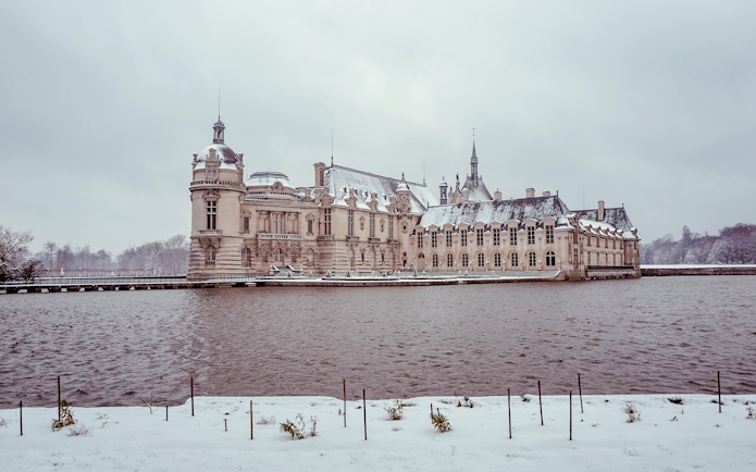 Chateau of Chantilly in winter with snow-covered grounds and surrounding water.