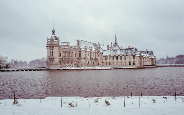 Chateau of Chantilly in winter with snow-covered grounds and surrounding water.