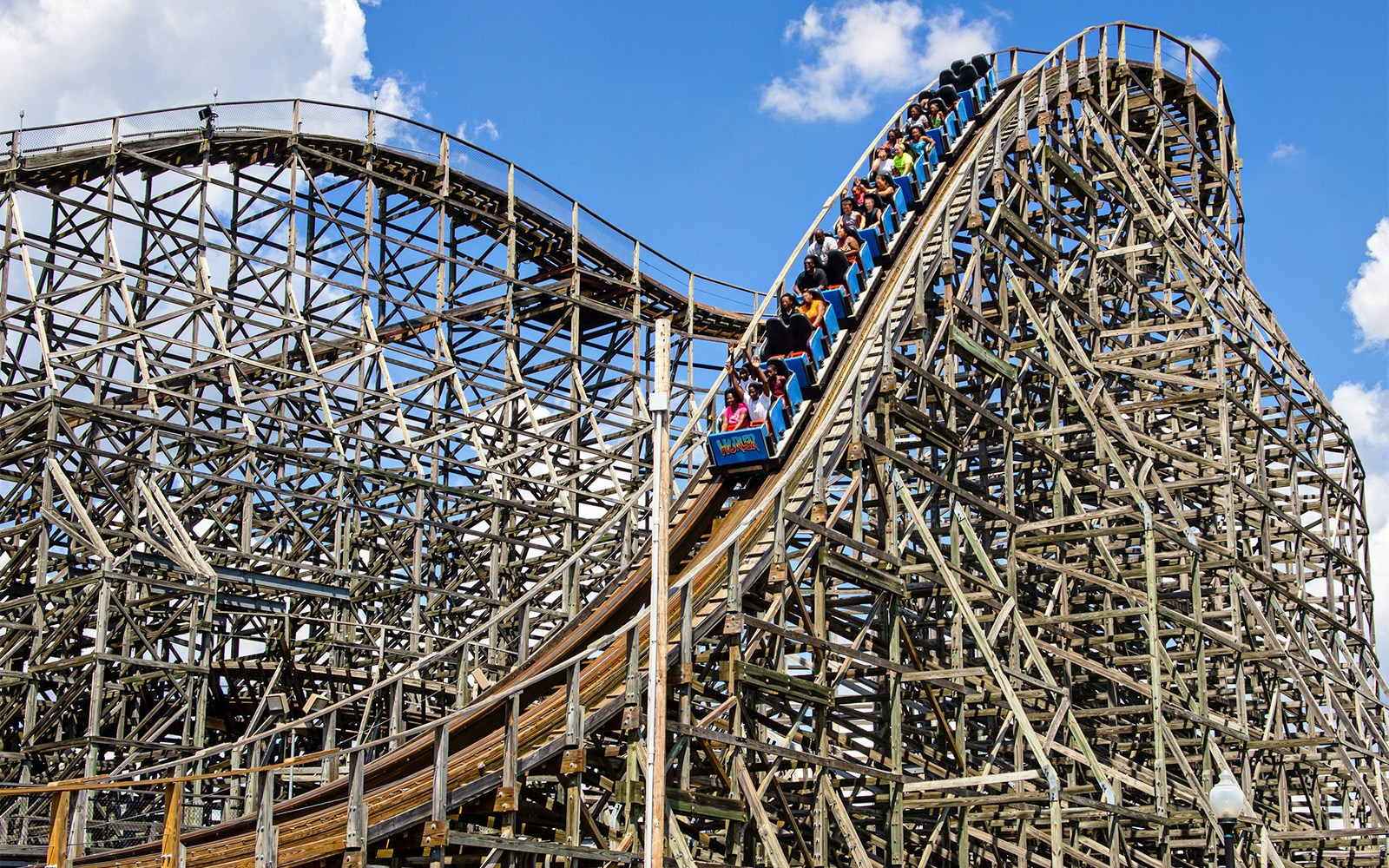 Wooden roller coaster with riders descending a steep track at a theme park during the day.