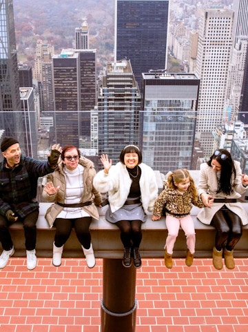 Visitors sitting on the edge at Top of the Rock, New York City skyline in the background.